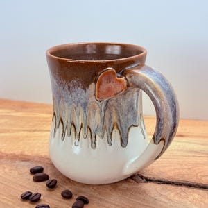 May include: A handmade ceramic mug with a heart-shaped accent on the handle. The mug features a brown rim, a white base, and a dripped glaze effect in shades of gray and brown. Coffee beans are scattered on the wooden surface.