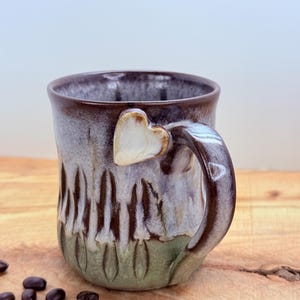 May include: A handmade ceramic mug with a heart-shaped handle attachment. The mug features a brown rim, a light blue and green body, and a textured design. Coffee beans are scattered on the wooden surface next to the mug.