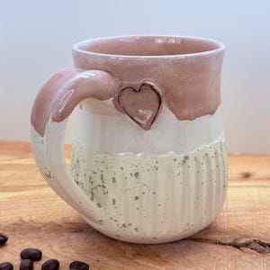 May include: A handmade ceramic mug with a heart-shaped accent. The mug features a pink rim, a white middle section with a speckled texture, and a ribbed bottom. The handle is pink and the heart is a bronze color.