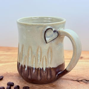 May include: A handmade ceramic mug with a heart-shaped accent on the handle. The mug features a two-tone design with a light beige upper half and a dark brown lower half. Coffee beans are scattered around the mug.