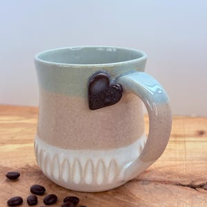 May include: A handmade ceramic mug with a heart-shaped handle. The mug features a color-blocked design with a light green top, tan middle, and white bottom with a textured pattern. Coffee beans are scattered on the wooden surface.