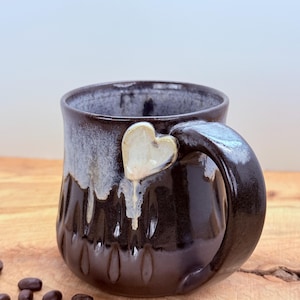 May include: A handmade ceramic mug with a dark brown glaze and a heart-shaped accent on the handle. The mug has a textured exterior and a dripped glaze effect. Coffee beans are scattered on the wooden surface.
