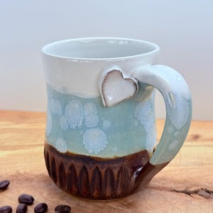 May include: A handmade ceramic mug with a heart-shaped handle attachment. The mug features a color gradient from white to light blue, with a textured brown base. Coffee beans are scattered on the wooden surface.