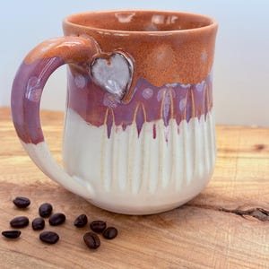 May include: A handmade ceramic mug with a heart-shaped accent on the handle. The mug features a gradient of colors, transitioning from a reddish-brown rim to a white base. Coffee beans are scattered on the wooden surface beside the mug.