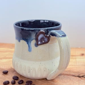 May include: Handmade ceramic mug with a heart-shaped handle attachment. The mug features a gradient of colors, including cream, beige, and navy blue. Coffee beans are scattered on the wooden surface near the mug. The mug is designed for hot beverages.
