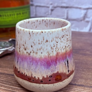 May include: A ceramic tumbler with a speckled cream-colored upper section, a vibrant purple and pink horizontal band in the middle, and a solid reddish-brown base. The surface features a reactive glaze finish with dark brown speckles throughout.