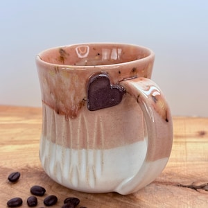 May include: A handmade ceramic mug with a heart-shaped accent on the handle. The mug features a color gradient from light pink to white, with a textured design. Coffee beans are scattered on the wooden surface.