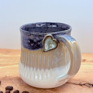 May include: A handmade ceramic mug with a heart-shaped handle. The mug features a gradient of colors, starting with a dark blue rim, transitioning to tan, and ending with a white base. Coffee beans are scattered on the wooden surface.