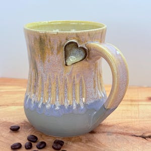 May include: A handmade ceramic mug with a heart-shaped accent on the handle. The mug features a textured design with a color gradient of yellow, pink, and blue. Coffee beans are scattered on the wooden surface next to the mug.