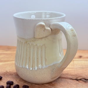 May include: Cream-colored ceramic mug with a heart-shaped accent on the handle. The mug has a textured, ribbed design on the lower half and a smooth, glossy finish on the upper half. Coffee beans are scattered on the wooden surface.