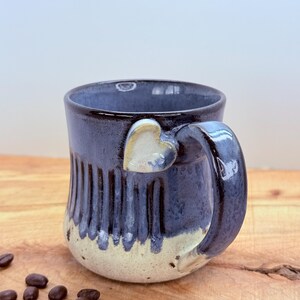 May include: A handmade ceramic mug with a navy blue glaze and a heart-shaped handle. The bottom of the mug is a cream color. Coffee beans are scattered on the wooden surface next to the mug.