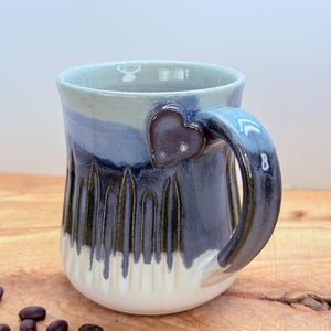 May include: A handmade ceramic mug with a heart-shaped accent on the handle. The mug features a gradient of colors, including blue, white, and a darker shade of blue. The mug has a unique textured design and is perfect for enjoying coffee or tea.