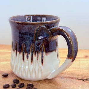 May include: A handmade ceramic coffee mug with a heart-shaped detail on the handle. The mug features a two-tone design, with a dark brown and blue upper section and a cream-colored, textured lower section. Coffee beans are scattered around the mug.