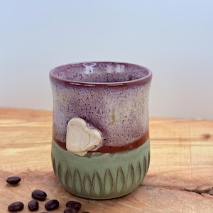 May include: A handmade ceramic mug with a heart-shaped embellishment. The mug features a color scheme of purple, beige, and green. Coffee beans are scattered on the wooden surface. The mug is designed for beverages.