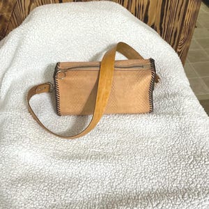 May include: A tan leather fanny pack with a textured snakeskin pattern. The bag has a long, adjustable strap and a zipper closure. The edges are stitched with black thread. The bag is sitting on a white, fluffy surface.