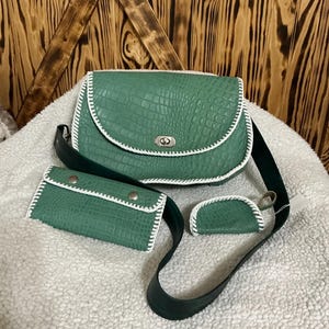 May include: A green faux-leather handbag set with a crocodile texture, white stitching, and a silver clasp. The set includes a crossbody bag, a wallet, and a small coin purse. The strap is dark green.