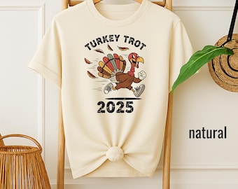 May include: Cream-colored t-shirt featuring a cartoon turkey running with the text "TURKEY TROT 2025". The turkey has a colorful fan of feathers and is wearing sneakers. The shirt is hanging on a wooden rack.