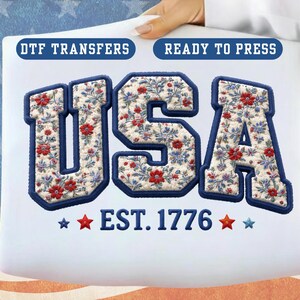 May include: A white DTF transfer with the word "USA" in a varsity-style font, filled with a floral pattern of red flowers and blue leaves. The text "EST. 1776" is below the letters, with red and blue stars. The words "DTF TRANSFERS" and "READY TO PRESS" are at the top.