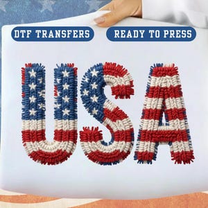 May include: A white fabric with the word "USA" in red, white, and blue, resembling the American flag. The letters are textured and have a stitched appearance. The text "DTF TRANSFERS" and "READY TO PRESS" are in blue boxes.