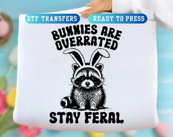 May include: White graphic featuring a raccoon wearing bunny ears, with the text "Bunnies Are Overrated Stay Feral." The design is in black and white, suitable for DTF transfers and ready to press.