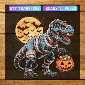 May include: A blue and pink cartoon dinosaur wrapped in bandages, holding a jack-o'-lantern bucket filled with candy. A full moon with bats and the text "DTF TRANSFERS READY TO PRESS" are in the background. The image is on a dark wood grain background.