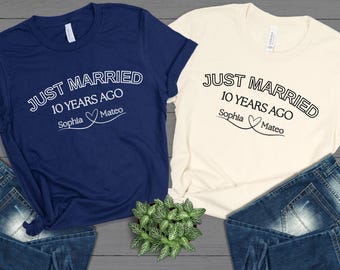 May include: Two t-shirts, one navy blue and one cream, with the text "JUST MARRIED 10 YEARS AGO" in white. The names "Sophia" and "Mateo" are below the text, connected by a heart. The shirts are paired with blue jeans.