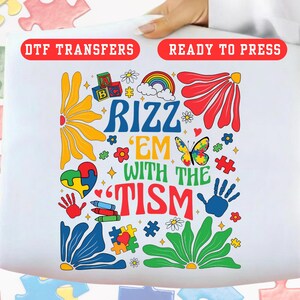 May include: White DTF transfer with the text "RIZZ 'EM WITH THE TISM" in blue, red, and green. The design includes a rainbow, puzzle pieces, flowers, a butterfly, and handprints. The words "DTF TRANSFERS" and "READY TO PRESS" are in red.
