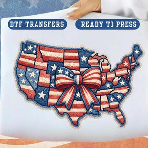May include: A white DTF transfer with a patriotic design. The design features a detailed embroidered map of the United States, incorporating the American flag's red, white, and blue colors. A large bow is centered over the map. The text "READY TO PRESS" is visible.