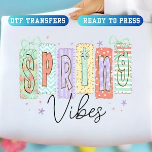 May include: A white DTF transfer with the words "SPRING VIBES" in colorful, decorative lettering. The word "SPRING" is in individual vertical rectangles with floral and patterned designs. The text "DTF TRANSFERS" and "READY TO PRESS" are in blue rectangles.