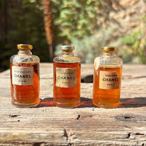 Puede incluir: Tres frascos de perfume Chanel vintage con tapas doradas y etiquetas. Las etiquetas dicen "Bois des Iles", "Cuir de Russie" y "Gardenia". Los frascos contienen líquido color ámbar y se exhiben sobre una superficie de madera envejecida.