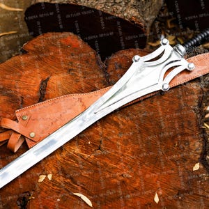 May include: A silver sword with a decorative hilt and a leather-wrapped handle rests on a wooden surface. The sword has a long, sharp blade and is accompanied by a brown leather sheath. The overall design suggests a historical or fantasy theme.
