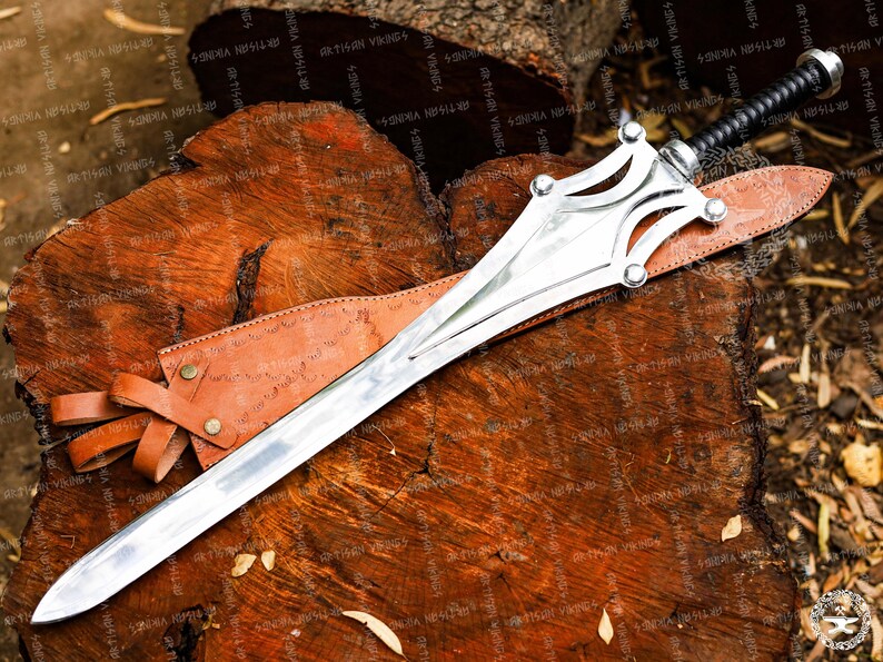 May include: A silver sword with a decorative hilt and a leather-wrapped handle rests on a wooden surface. The sword has a long, sharp blade and is accompanied by a brown leather sheath. The overall design suggests a historical or fantasy theme.