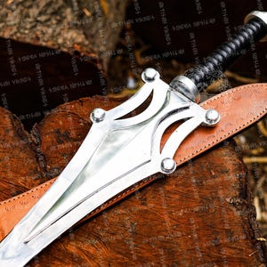May include: A silver sword with a decorative hilt and a black wrapped handle rests on a brown leather sheath. The sword is displayed on a wooden surface, showcasing its intricate design and craftsmanship.