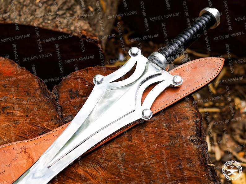 May include: A silver sword with a decorative hilt and a black wrapped handle rests on a brown leather sheath. The sword is displayed on a wooden surface, showcasing its intricate design and craftsmanship.