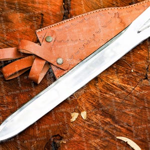 May include: A polished silver sword with a leather sheath and handle, resting on a textured wooden surface. The sheath is brown with decorative patterns and secured with a leather strap. The sword's blade is long and sharp.