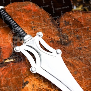 May include: A silver-colored sword with a black, textured handle. The blade is wide and flat, with a decorative guard featuring rounded accents. The sword rests on a textured, brown wooden surface.