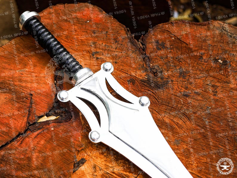 May include: A silver-colored sword with a black, textured handle. The blade is wide and flat, with a decorative guard featuring rounded accents. The sword rests on a textured, brown wooden surface.