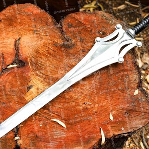 May include: A silver-colored sword with a decorative hilt and a black handle rests on a wooden surface. The blade is long and pointed, with a central ridge. The hilt features a stylized design with rounded accents. The handle is wrapped in a textured, black material.