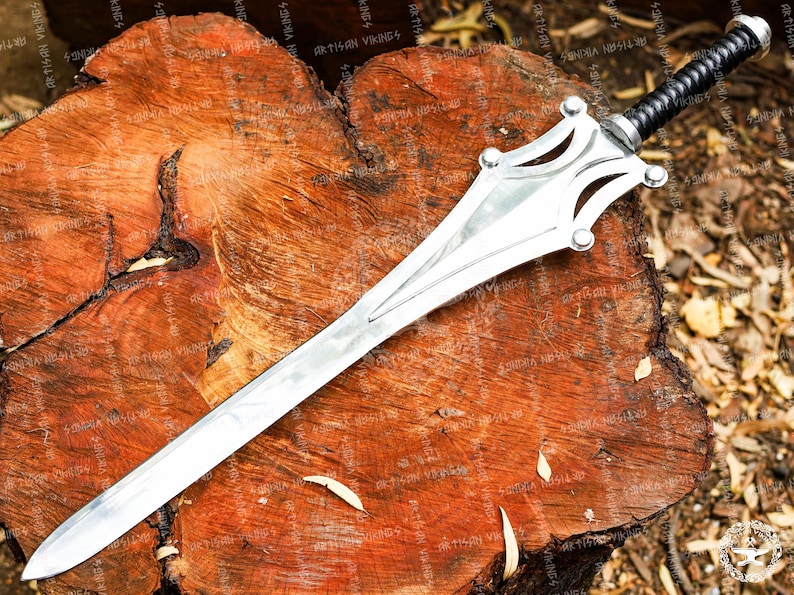 May include: A silver-colored sword with a decorative hilt and a black handle rests on a wooden surface. The blade is long and pointed, with a central ridge. The hilt features a stylized design with rounded accents. The handle is wrapped in a textured, black material.