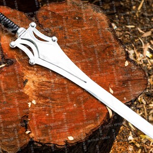 May include: A silver sword with a black wrapped handle, resting on a weathered, brown wooden surface. The sword features a distinctive, stylized hilt design with rounded accents. The blade is long and straight, reflecting the light.