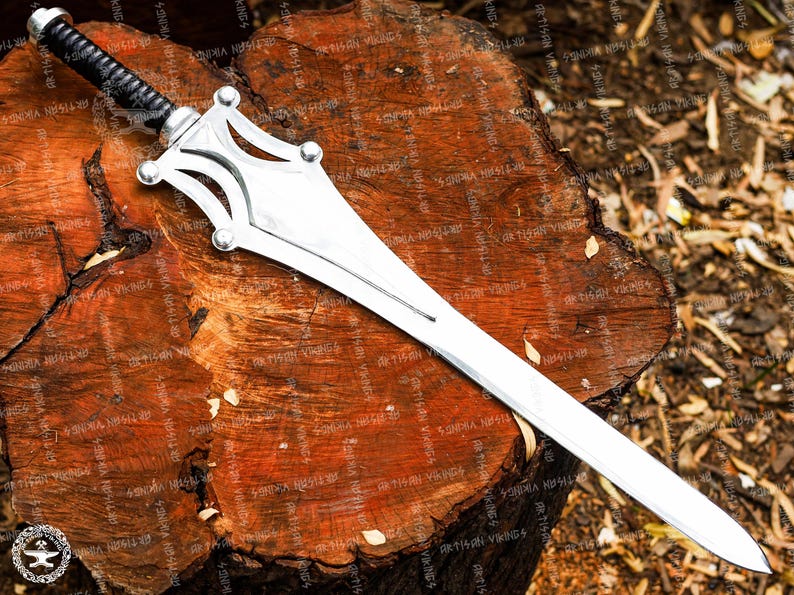 May include: A silver sword with a black wrapped handle, resting on a weathered, brown wooden surface. The sword features a distinctive, stylized hilt design with rounded accents. The blade is long and straight, reflecting the light.