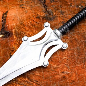 May include: A silver sword with a black leather-wrapped handle. The blade is polished and has a decorative guard with rounded accents. The sword is set against a rustic, wooden background.