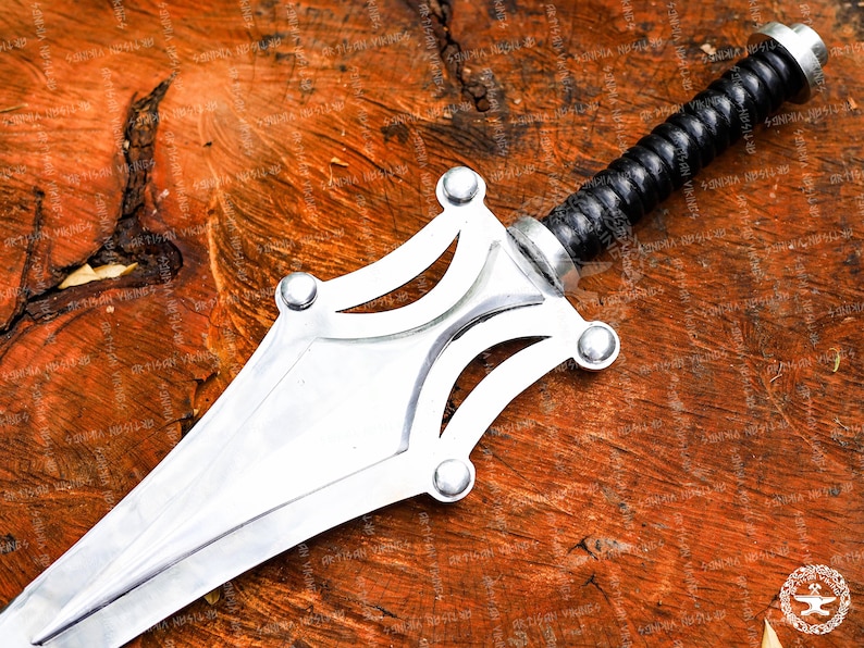 May include: A silver sword with a black leather-wrapped handle. The blade is polished and has a decorative guard with rounded accents. The sword is set against a rustic, wooden background.