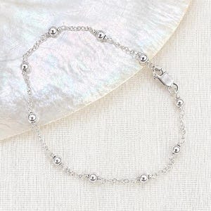 May include: A delicate silver chain bracelet featuring evenly spaced small polished silver beads throughout the length of the chain. The bracelet includes a secure lobster claw clasp mechanism for fastening, creating a simple and refined accessory.