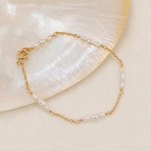Op de afbeelding: Een verfijnde goudkleurige kettingarmband met kleine, witte, ovaalvormige zoetwaterparels die gelijkmatig over de schakels zijn verdeeld. De armband is voorzien van een ronde veerringsluiting en heeft een elegant ontwerp met afwisselend metaal.