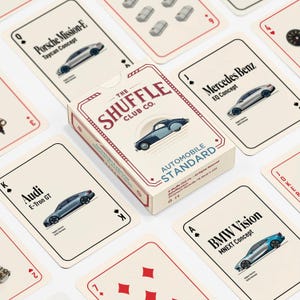 May include: A deck of playing cards with a box featuring the text "The Shuffle Club Co." and "Automobile Standard." The cards display car illustrations, including Audi, Porsche, and BMW models. The cards are arranged on a white surface.