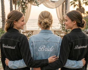 May include: Three people wearing denim jackets with embroidered text. One jacket is light blue with "Bride" embroidered on the back. The other two jackets are black with "Bridesmaid" embroidered on the back. The jackets are worn in front of a beach wedding setting.