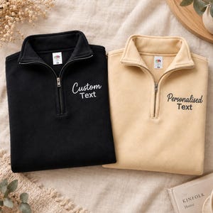 May include: A black quarter-zip sweatshirt with the embroidered text Custom Text and a beige quarter-zip sweatshirt with the embroidered text Personalised Text. Both garments feature a high collar, a metal zip, and a Fruit of the Loom label.