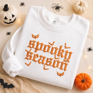May include: A white crewneck sweatshirt featuring orange embroidered text that reads Spooky Season. The design includes small orange bat motifs surrounding the lettering. The garment has long sleeves and a ribbed collar, cuffs, and hem.