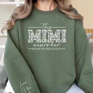 以下が含まれることがあります： オリーブグリーンのスウェットシャツ。白で「this MIMI wears her HEART ON HER SLEEVE」と書かれています。「MIMI」の文字はヒョウ柄のデザインです。袖には「Luna Ma」とハートの文字が入っています。快適でスタイリッシュなアパレルアイテムです。